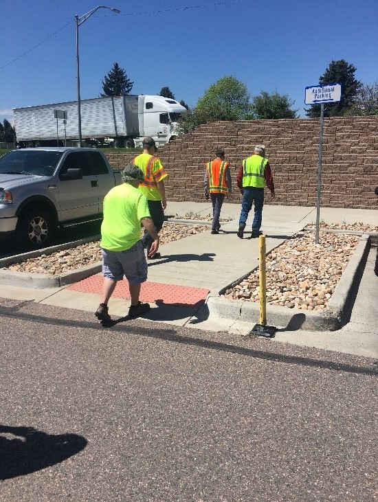 Four individuals in yellow vests wearing vision impairment goggles crossing at a curb ramp. 