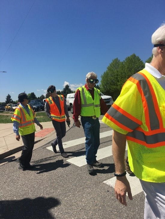 Four men in yellow safety vests and visual impairment goggles crossing an intersection at a crosswalk. 