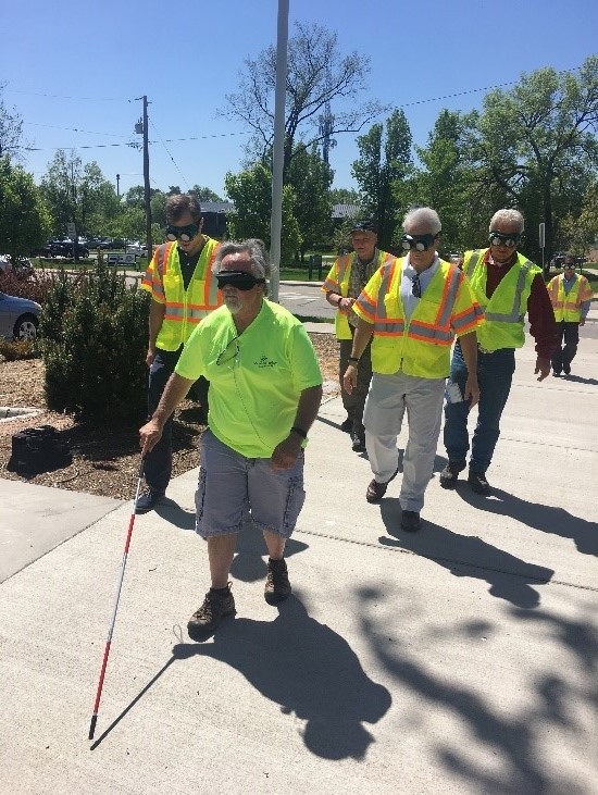 Six men in yellow vests and visual impairment goggles walking on a sidewalk. The individual in the front is also using a white cane. 