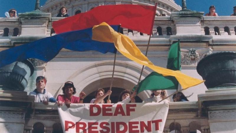 Several people in front of a white building with colorful flags and a sign reading "Deaf President Now"