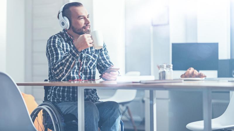Bearded man sips coffee at a café while listening to music on his headphones while sitting in his wheelchair.