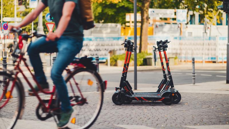 E-scooters are parked on a sidewalk as someone on a bike rides past.