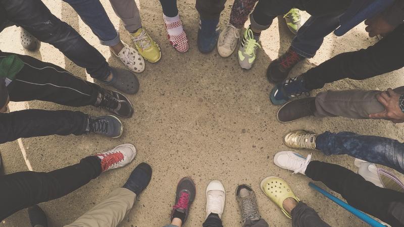 A circle is formed from a group of people all putting a foot down standing next to one another. Only the lower leg and shoes are pictured.