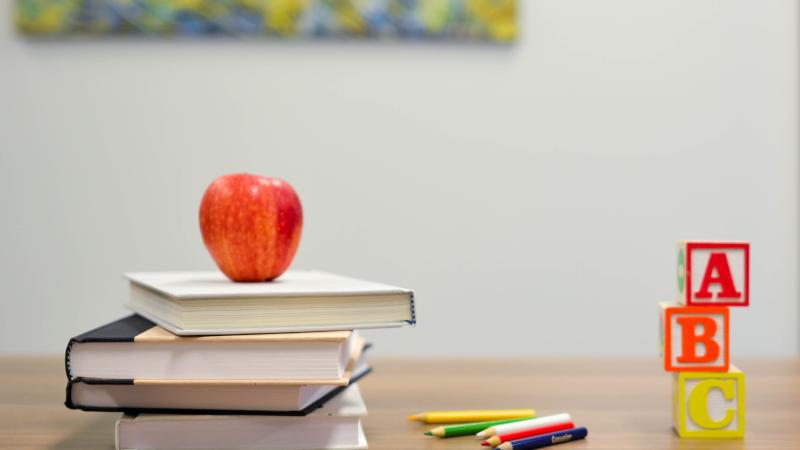 A red apple on top of 3 books next to colored pencils and alphabet blocks.