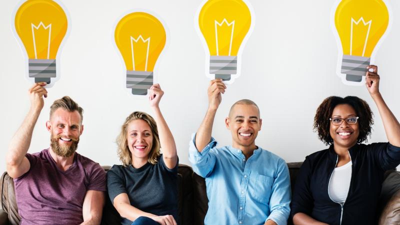 Four smiling people sitting on a couch holding cutouts of light bulbs above their heads to symbolize ideas.