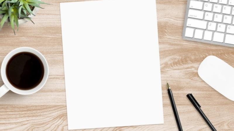 A blank piece of paper and pens on a desk, with a keyboard and a cup of coffee.