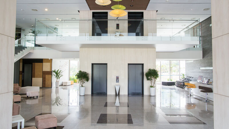 The interior of a wide space in a public building. Centered are 2 elevator shafts to the second floor with a reflective title flooring. Kiosks and seating area frame the sides with moderately sized planters peppered throughout.