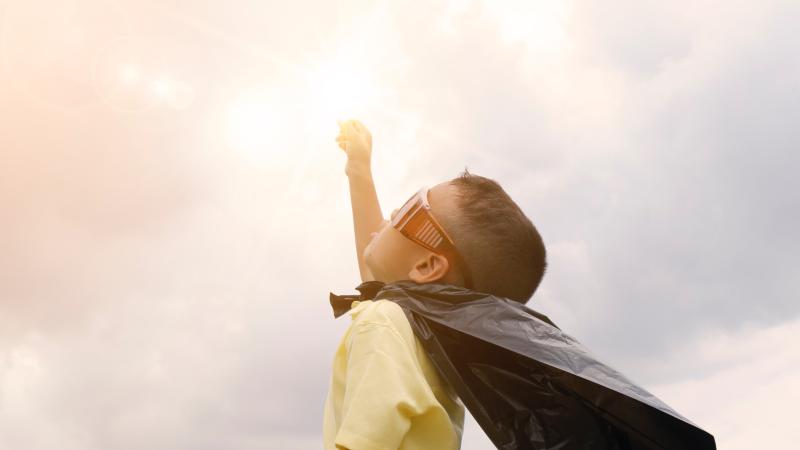 A small boy is dressed as a super hero with a make-shift cape and wide sunglasses and is reaching towards the sky with one hand, intersecting the sun in the background.