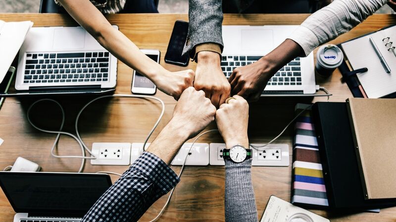 A 5-person fist bump, above a desk with several laptop computers and note pads.