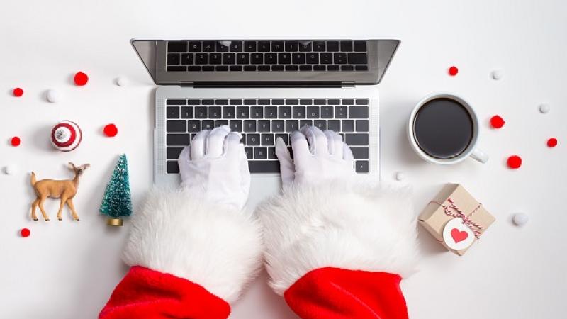 A man wearing a read suit and white gloves typing on a laptop, sitting on a desk full of holiday items.