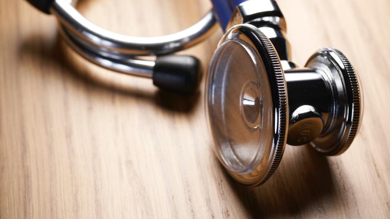 A doctor’s stethoscope is laying on a wooden table, with the chest piece laying over the earpieces.