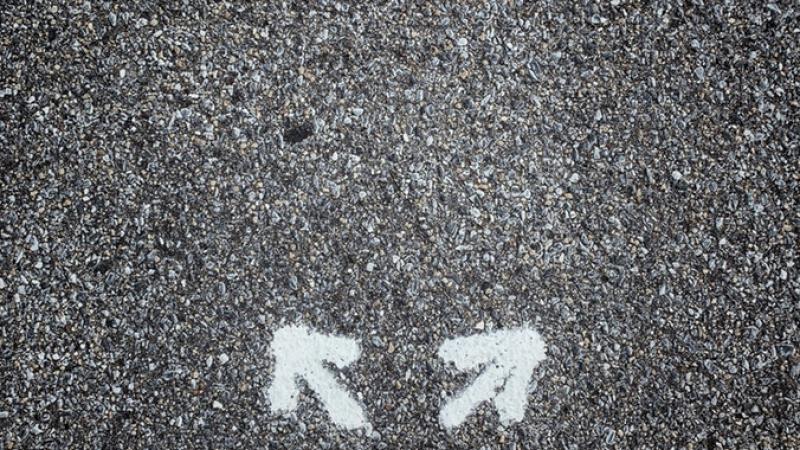 A photo of the ground with the photographer’s dress shoes in view in front of a pair of arrows painted on the asphalt, one point forward-right, the other pointing forward-left.
