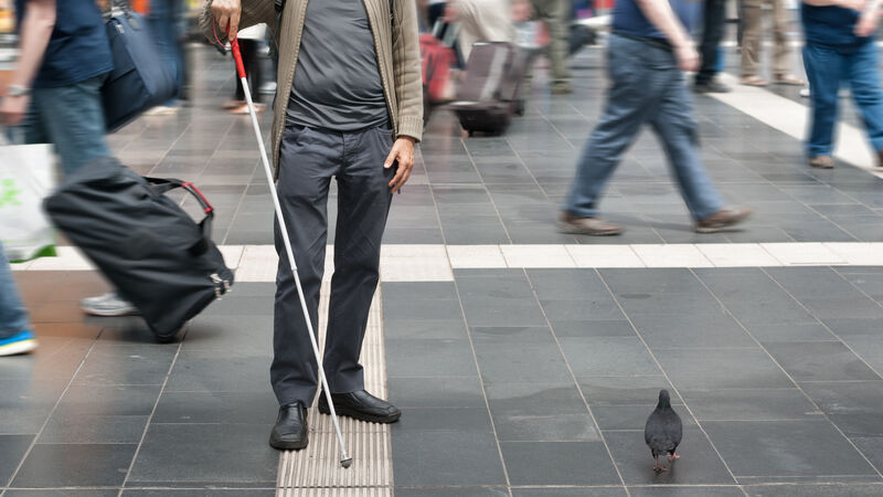 A blind man is navigating through a busy airport using a white cane to follow a textured strip on the ground.