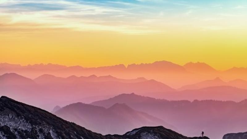 A landscape photograph of the colorful sunset at top of a mountain, with a hiker in the background.