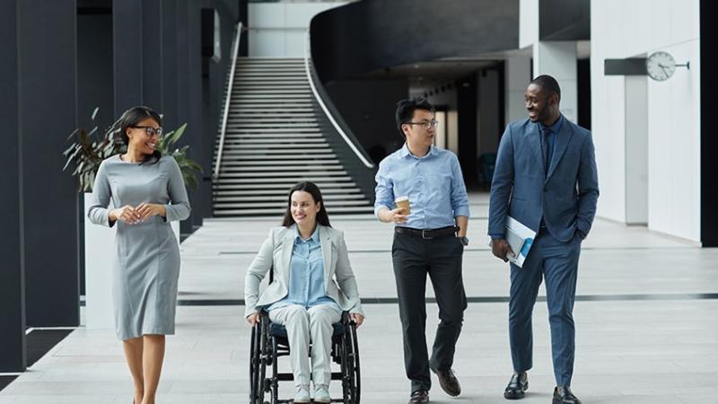A photograph of a group of diverse coworkers, including a person in a wheelchair, walking and having a conversation in the lobby of a building.