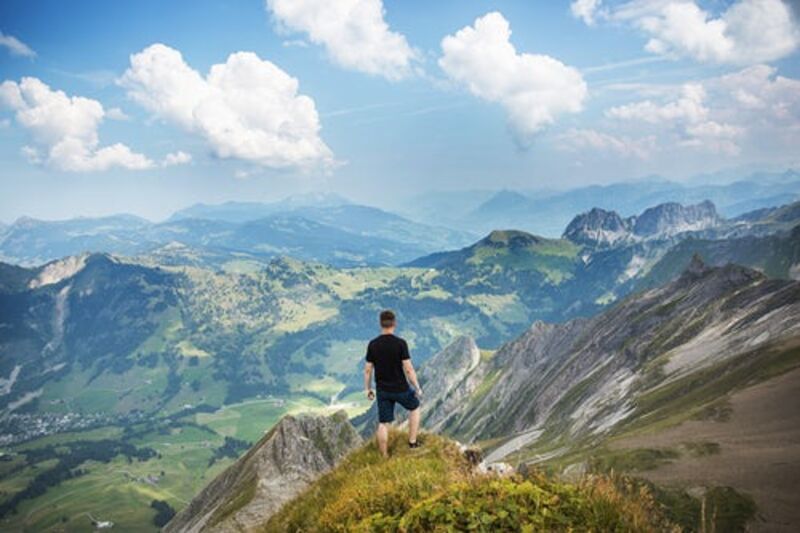 Hiker standing on a grassy peak over looking a vast valley below with mountains in the distance on a sunny day.