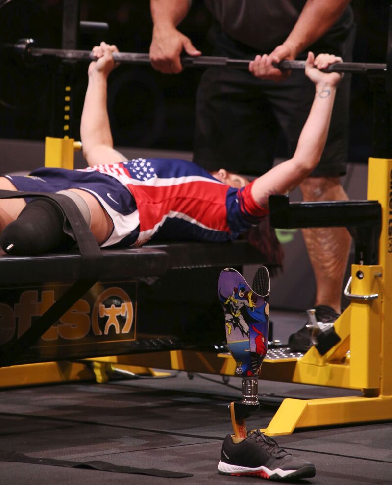 A below the knee amputee is competing in  power lifting bench press, with a spotter ensuring safety and the prosthetic lower limb off the leg, standing by itself in the foreground.