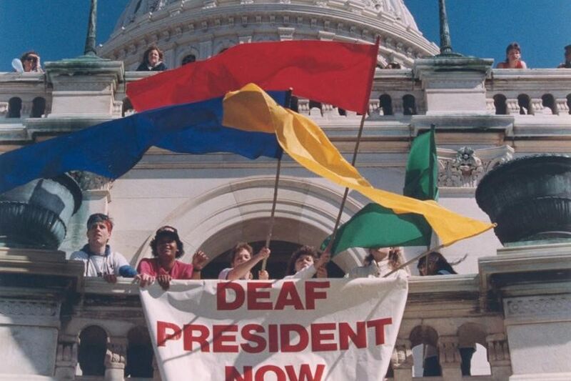 Several people in front of a white building with colorful flags and a sign reading "Deaf President Now"