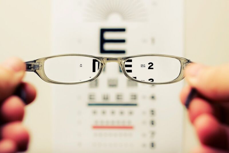 In the foreground, a pair of glasses held up to a blurry visual acuity chart, showing smaller and smaller letters. The glasses bring the poster into focus.