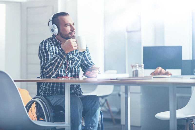 Bearded man sips coffee at a café while listening to music on his headphones while sitting in his wheelchair.