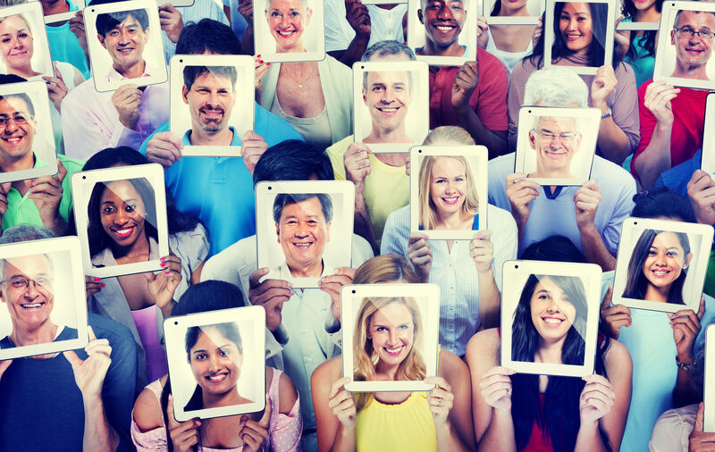 A colorful photo of around 2-dozen people of all shapes and sizes all holding an electronic table in front of their faces with a photo of their faces smiling on the screens.