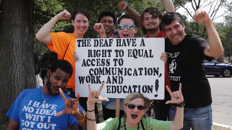 Group of people holding a sign that says THE DEAF HAVE A RIGHT TO EQUAL ACCESS TO COMMUNICATION WORK AND EDUCATION!