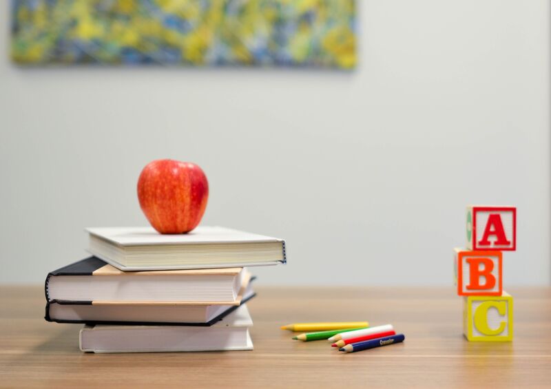 A red apple on top of 3 books next to colored pencils and alphabet blocks.