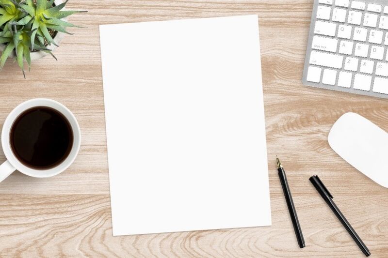 A blank piece of paper and pens on a desk, with a keyboard and a cup of coffee.