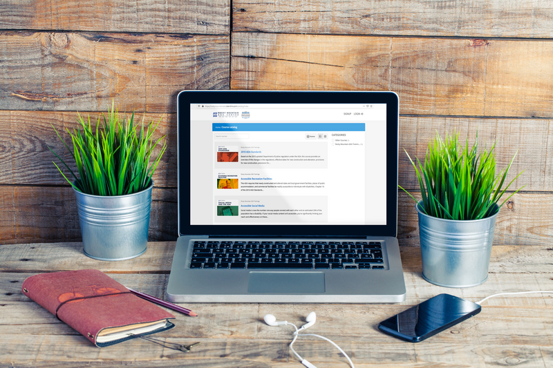 A laptop with the RMADAC online portal page on screen, sitting on a wooden desk with two plants, a notebook, and a cellphone.