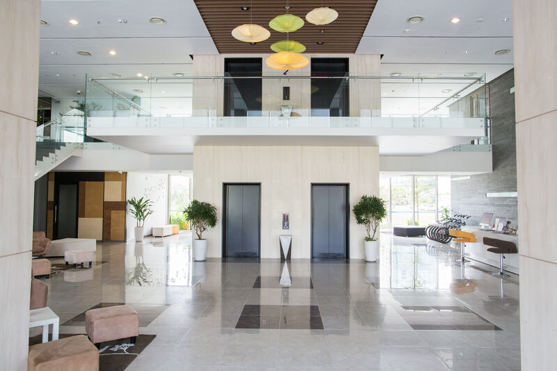 The interior of a wide space in a public building. Centered are 2 elevator shafts to the second floor with a reflective title flooring. Kiosks and seating area frame the sides with moderately sized planters peppered throughout.