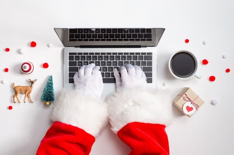 A man wearing a read suit and white gloves typing on a laptop, sitting on a desk full of holiday items.