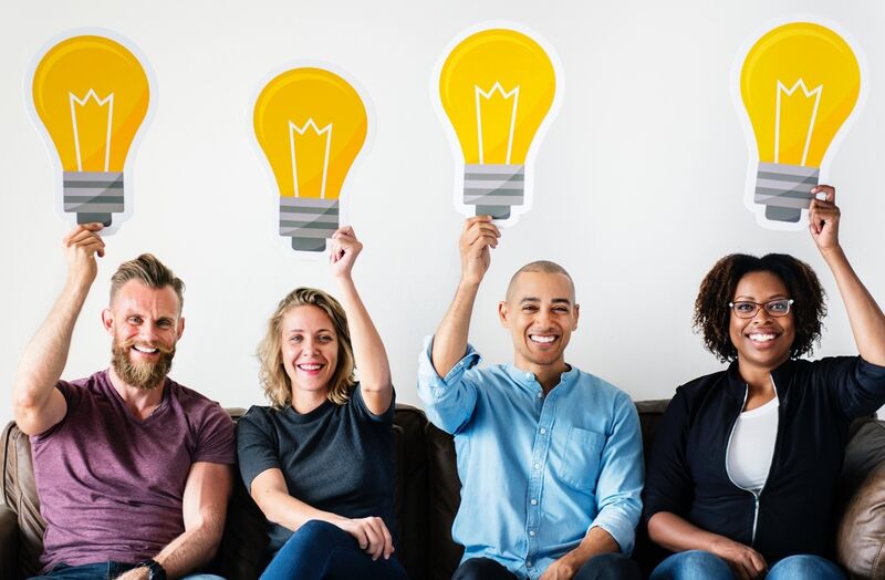 Four people sitting on a couch smiling holding pictures of light bulbs above their heads.