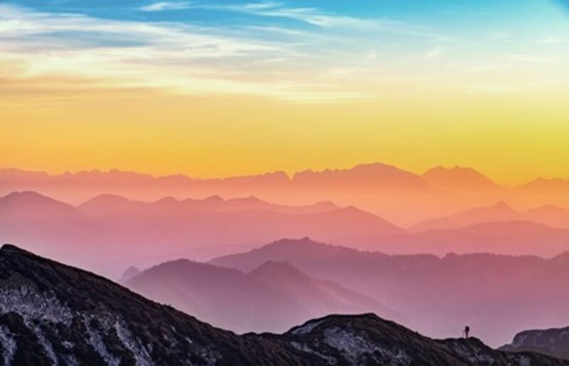A landscape photograph of the colorful sunset at top of a mountain, with a hiker in the background.