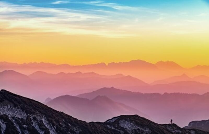 A landscape photograph of the colorful sunset at top of a mountain, with a hiker in the background. 