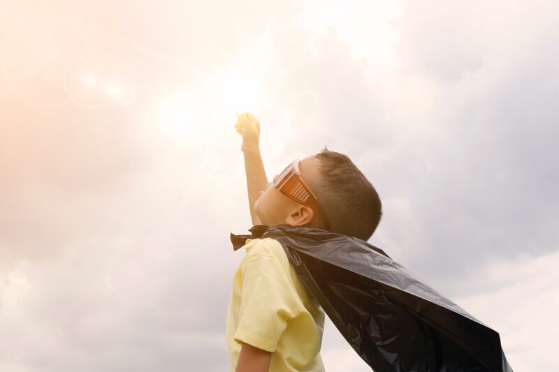 A little boy dressed like a superhero, wearing a cape and goggles, holds a fist towards the sun in a cloudy sky.