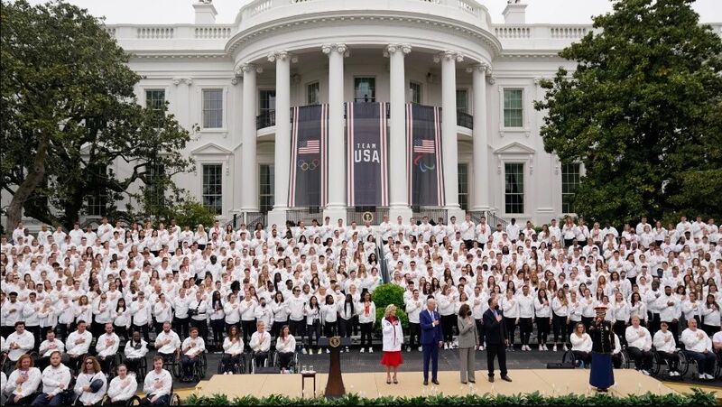 a group photo of Team USA athletes with President Joe Biden, Vice President Kamala Harris, and their spouses standing in front of the White House as a Marine sings the National Anthem