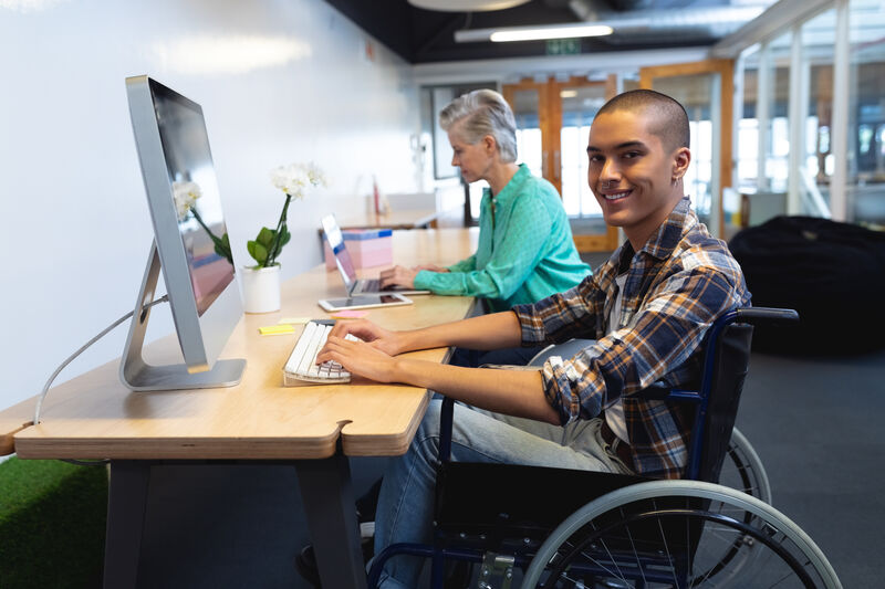 young Hispanic man sitting in his wheelchair smiling towards the camera, sitting at the computer desk