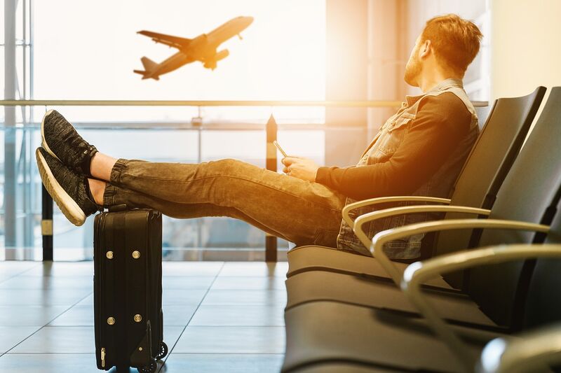 a young man gazing out the window at an airplane, sitting on a chair at the airport with his legs on his suitcase