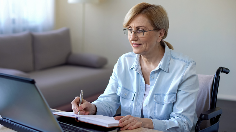 a senior woman in a wheelchair studying online and taking notes