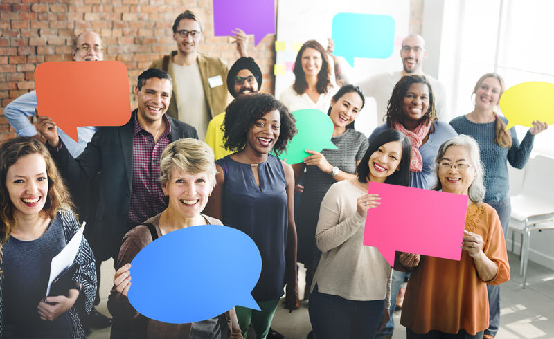 a crowd of people of all skin colors smiling at the camera, a few of them holding different colored thinking bubbles