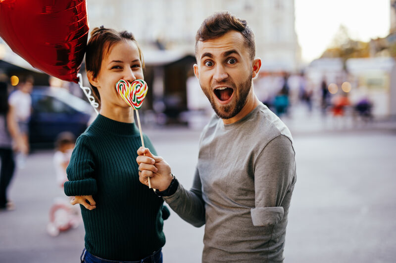 a young straight couple pose for a photo, both with upper limb disabilities. the male holds a heart-shaped lollipop in front of the female's mouth, she is smiling with her eyes and holding a red heart-shaped balloon.