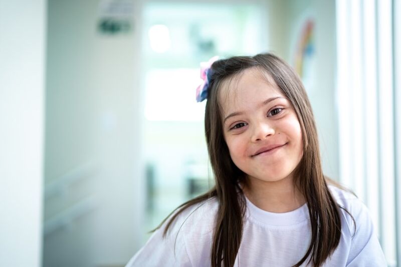 young brown-haired girl with Down syndrome smiling at the camera in the hallway of her school