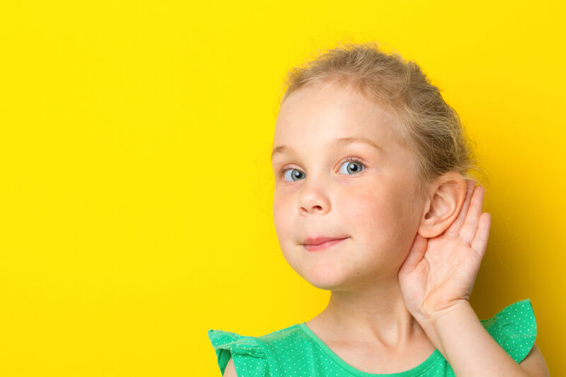 a young white blonde-haired girl standing in front of a yellow background, holding her ear as if she is trying to listen