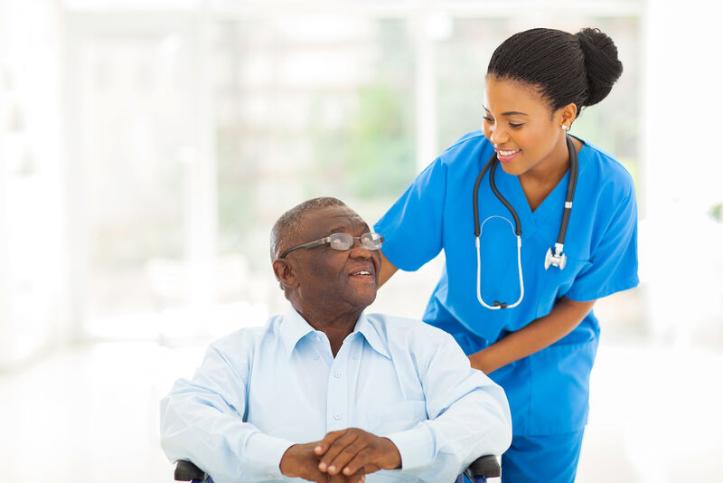 A nurse wearing blue scrubs with a stethoscope around her neck is pushing an older adult male patient in a wheelchair, while looking at him and smiling.
