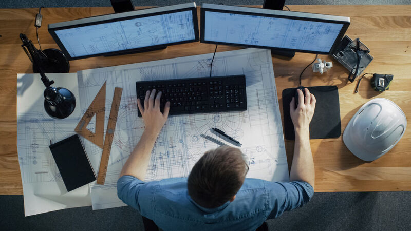 an overhead shot of an architect sitting at a draft table using computer software with two monitors and a printed schematic to work on a design