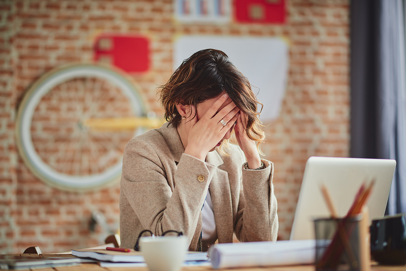 woman holds her head with her hands as she sits at her work desk