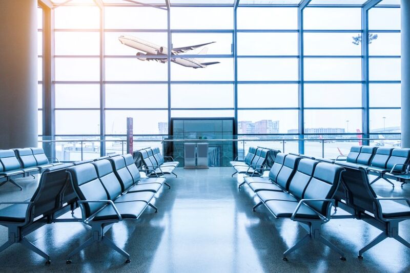 a photo of empty rows of black chairs at an airport terminal and windows and a plane taking off on a sunny day