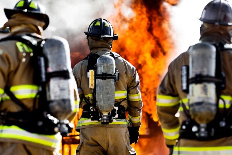 a fire rages in the distance as 3 fire fighters look upon the blaze, backs facing the camera