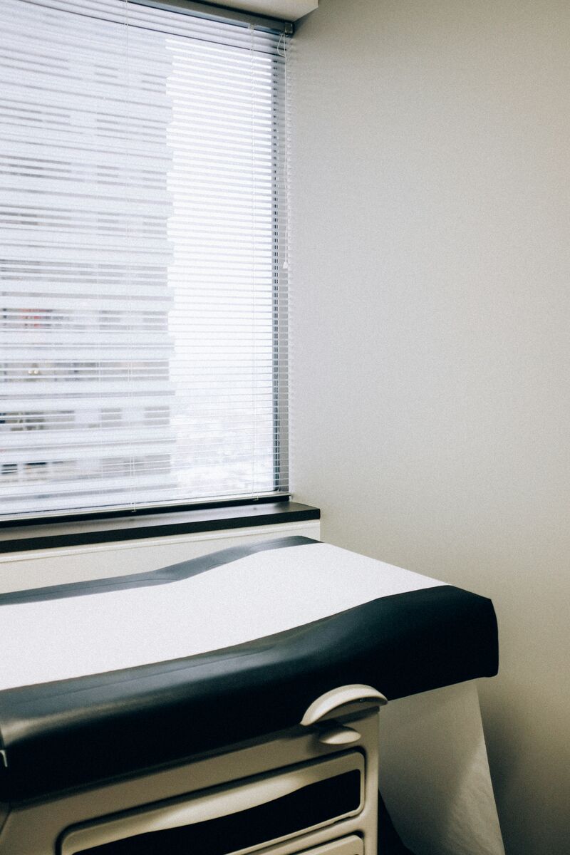 A medical examination table is near a window in a doctor’s office, with paper on top of the table and storage drawers underneath the top of the table.  