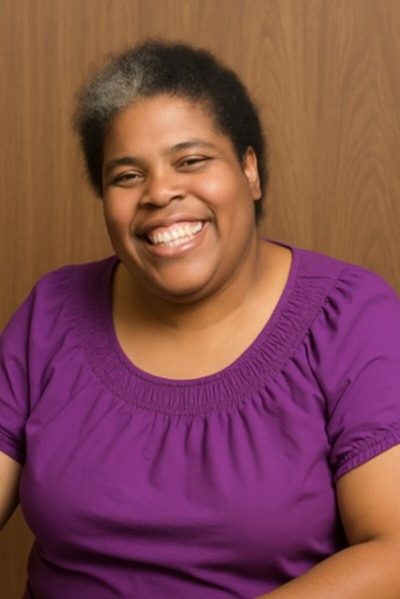 a black woman with short black hair that has a white spot smiling at the camera, wearing a purple shirt on a light brown background